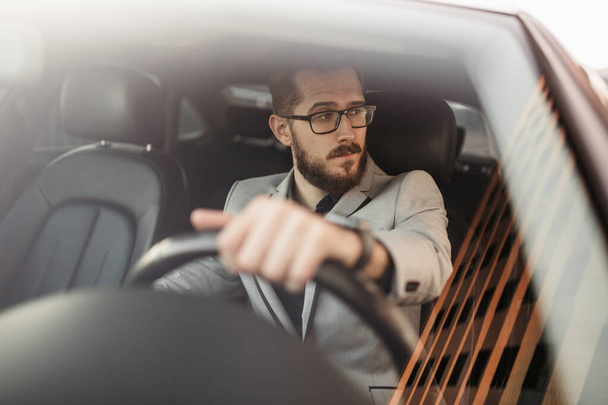 A bearded businessman with glasses sits behind the wheel of an Executive car and looks away. Photo through the windshield - Foto, Bild