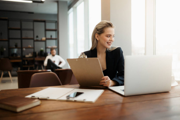 Egy közgazdász lány klasszikus öltönyben ül egy asztalnál egy laptoppal a munkahelyén, és mosolyogva néz ki az ablakon. - Fotó, kép