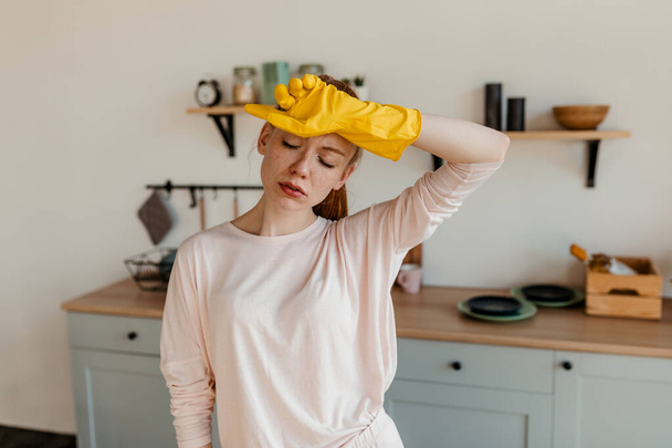 Red-haired girl in rubber gloves, standing in the kitchen, and suffering from a headache - Foto, immagini