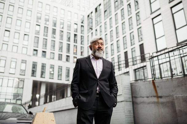 A bearded elderly businessman stands in the background of the business center and looks ahead - Photo, image