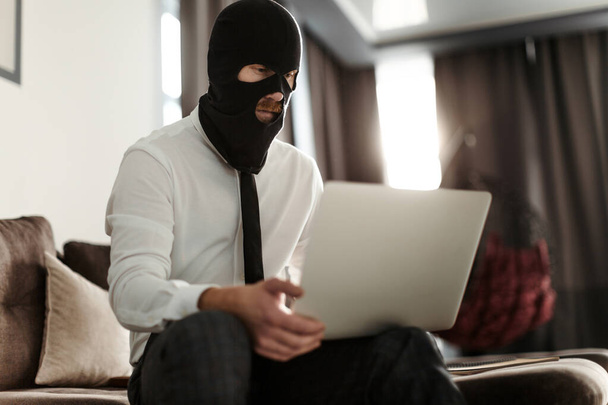 A man in a Balaclava mask sits at a laptop and publishes fake news - Foto, Imagem