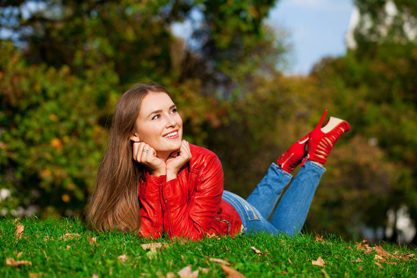 Young beautiful woman in red leather jacket and blue jeans sitting in the autumn park - Foto, Bild