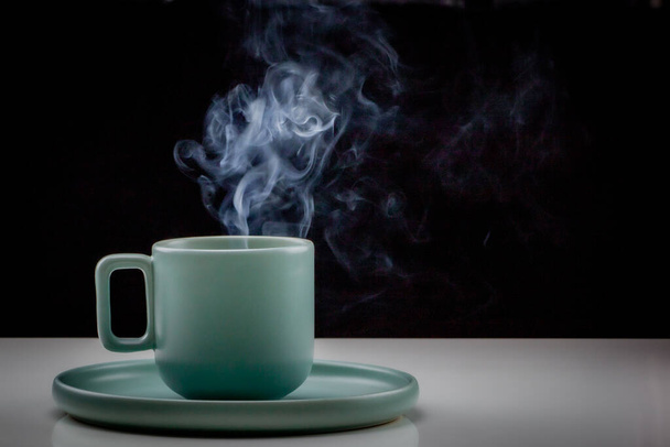 Pastel groen gekleurde koffie of thee kopje met hete vloeistof, rook en stoom, zwarte achtergrond. Beker staande op een licht reflecterende tafel die vervaagt tot een zwarte achtergrond - Foto, afbeelding