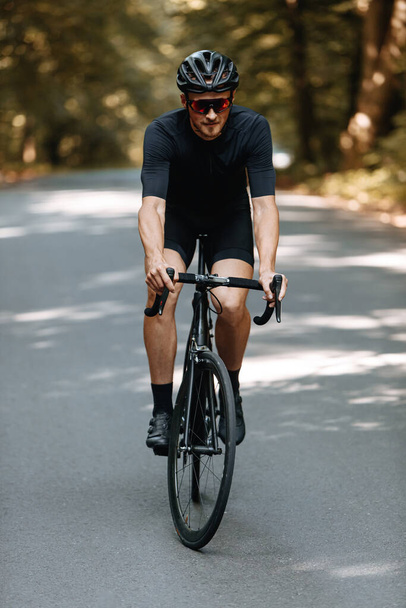 Elülső nézet erős férfi kerékpáros sportruházat lovaglás fekete kerékpár homályos háttér. Szakállas férfi védő sisak és szemüveg javítása készségek és felkészülés a versenyre - Fotó, kép