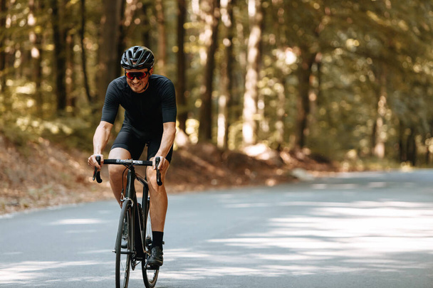 Happy bearded person in protective helmet, mirrored glasses and activewear biking on paved road in green forest during summer days. Concept of regular training and healthy lifestyle. - Photo, Image