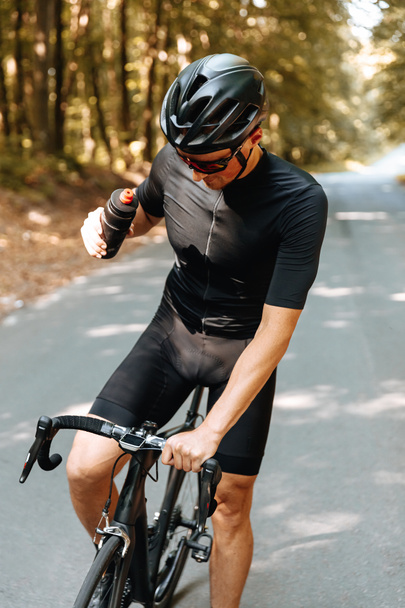 Professionele fietser in beschermende helm en bril gestopt in het groene bos om vers water te drinken. Volwassen man in activewear rust nemen tijdens het rijden. - Foto, afbeelding