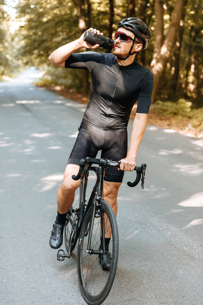 Ganzkörperporträt eines reifen bärtigen Mannes in Sportbekleidung, der sich nach langen Radtouren mit Wasser erfrischt. Starker Mann mit Helm und Brille trinkt frisches Getränk aus Sportflasche. - Foto, Bild