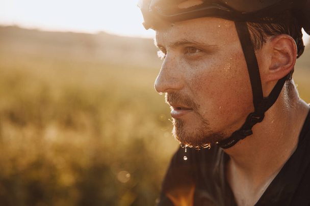 Egy fekete sisakos szakállas férfi arcképe, aki a szabadban biciklizés miatt fáradtnak érzi magát. Erős kerékpáros, akinek izzadságcseppek vannak az arcán, miközben edzés közben szünetet tart.. - Fotó, kép