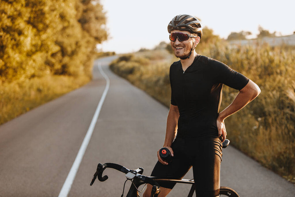 Vrolijke man met baard in activewear, zwarte helm en sportbril zittend op de fiets en kijkend op de camera met prachtige natuur rondom. Concept van een actieve en gezonde levensstijl. - Foto, afbeelding