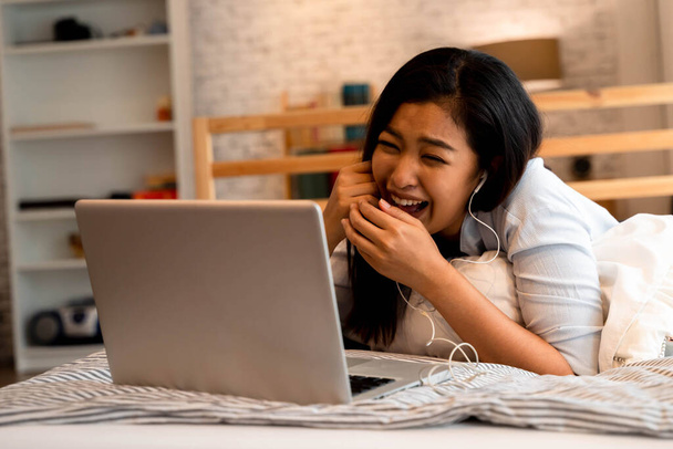 Portrait of happy young Asian girl in casual clothing lying down on bed while making a video call with laptop computer in bedroom at home. Video conferencing technology concept - 写真・画像