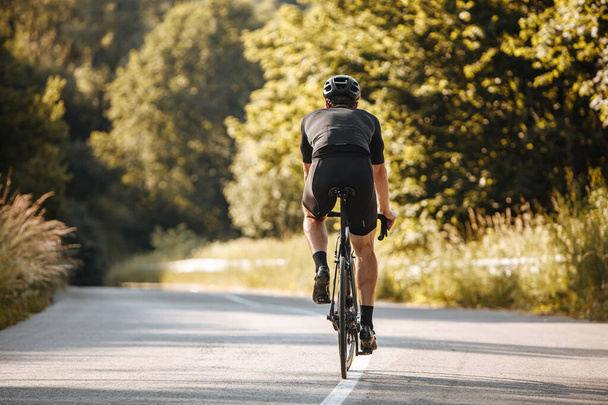スポーツウェアや黒のヘルメットでの強い男のバックビューは、自然上のプロの自転車に乗る。舗装道路で好きな趣味を楽しむ熟女スポーツマン. - 写真・画像