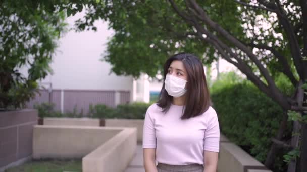Mladý dospělý asijské žena nosit bílou masku chůze k fotoaparátu na venkovní park chodník pod stromy odstíny, sám domácí karanténa, covid19 corona virus, přední pohled, 1080p zpomalení pocit osamělosti - Záběry, video