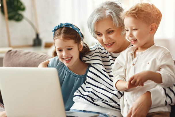 Веселая пожилая женщина наслаждается временем с маленькими внуками, сидя вместе на диване и смотря смешную карикатуру на лапто
 - Фото, изображение