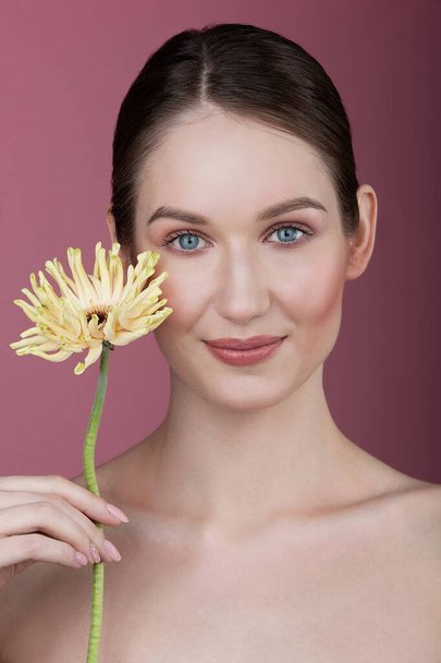 portrait of a girl with a flower on a pink background, natural makeup and even skin on her face, a smile and expensive eyes - Foto, Bild