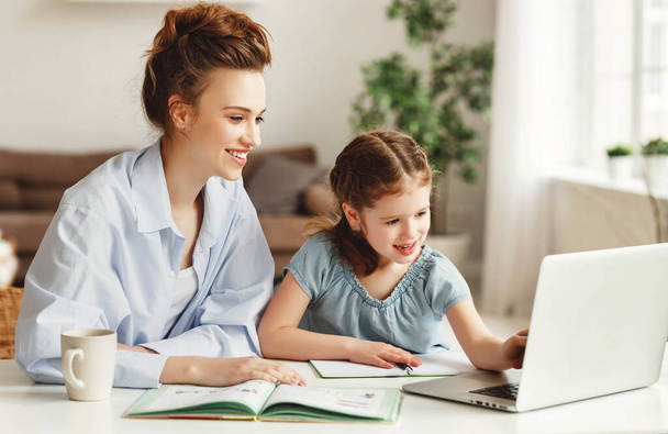 Pozitivní mladá žena pomáhá dceři při hledání informací pro domácí úkoly na internetu, zatímco sedí společně u stolu s notebookem na hom - Fotografie, Obrázek