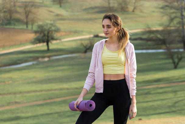 Una joven chica atlética delgada en ropa deportiva realiza un conjunto de ejercicios. Fitness y estilo de vida saludable en el fondo de verdes colinas de pastos de primavera. - Foto, Imagen
