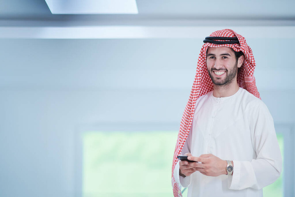молодой современный мусульманский арабский бизнесмен в традиционной одежде, используя смартфон дома - Фото, изображение