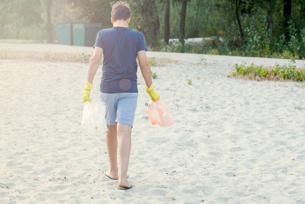 Niño en camiseta blanca con guantes recoge la basura y botellas de plástico en el paquete en la playa. Joven voluntario. Protección del medio ambiente, ahorrar concepto de medio ambiente
 - Foto, Imagen
