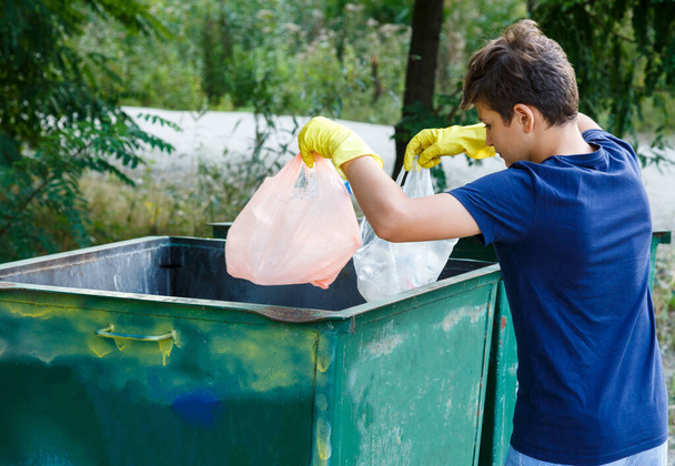 Eldivenli mavi tişörtlü çocuk çöpleri ve plastik şişeleri plajda paketlere doldurup konteynıra atıyor. Genç gönüllü. Çevre koruma, çevre konseptini kaydet - Fotoğraf, Görsel