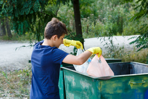 Junge in blauem T-Shirt mit Handschuhen sammelt Müll und Plastikflaschen in Paketen am Strand und wirft sie in Container. Junge Freiwillige. Umweltschutz, Umweltschutzkonzept - Foto, Bild