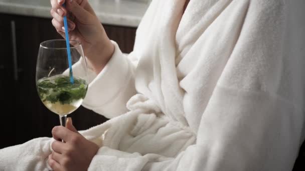 Girl is drinking mojito. Woman in kitchen with towel on head and in bathrobe drinks alcoholic mojito and looks out window. Close-up - Πλάνα, βίντεο