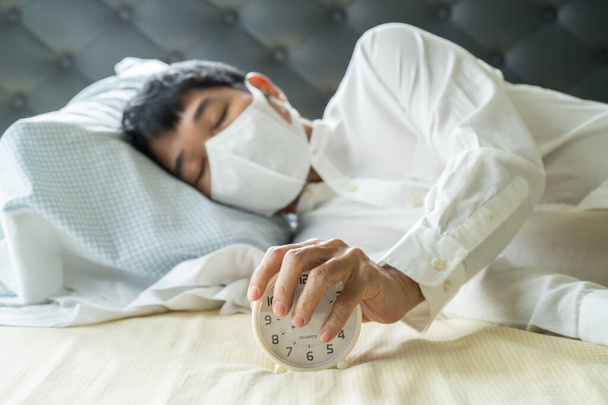 目覚まし時計でベッドの上で眠って外科マスクを身に着けているアジアのビジネスマン - 写真・画像