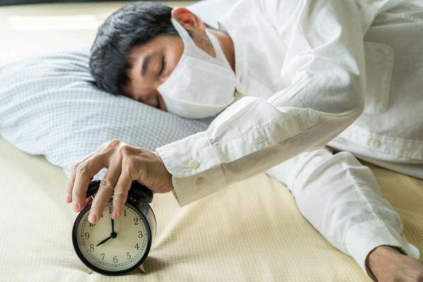 Азиатский бизнесмен в хирургической маске спит на кровати с будильником
 - Фото, изображение