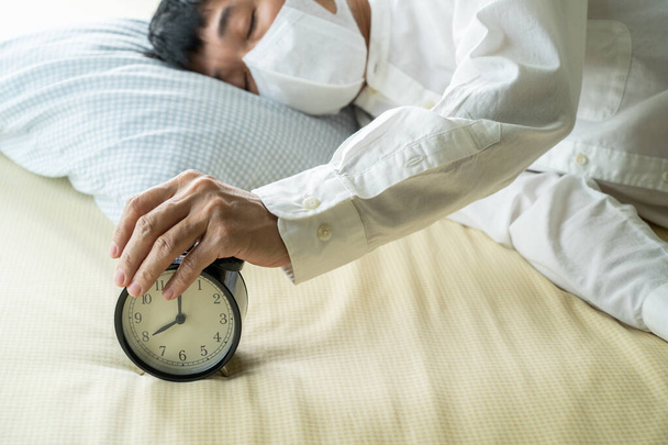 Азіатський бізнесмен у хірургічній масці спить на ліжку з будильником. - Фото, зображення