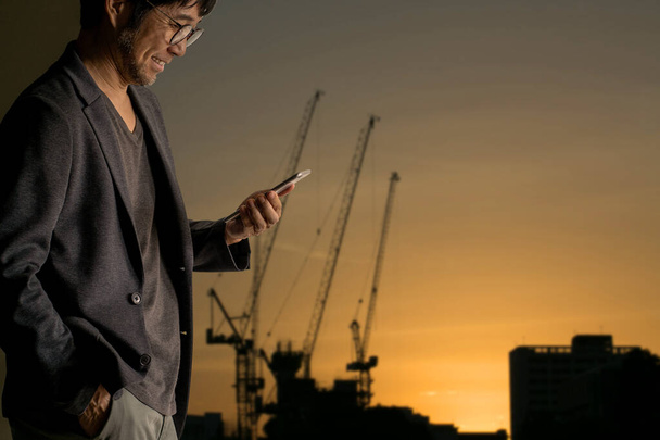 Азиатский бизнесмен держит смартфон с размытой строительной площадкой на закате
 - Фото, изображение