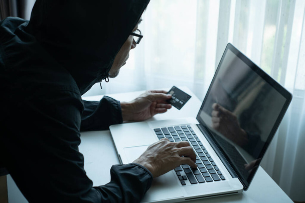 Πίσω μέρος του άνδρα ντύσιμο μαύρο σακάκι κουκούλα κρατώντας πιστωτική κάρτα και τον έλεγχο του κωδικού πρόσβασης στον υπολογιστή labtop. έννοια scammer χάκερ - Φωτογραφία, εικόνα