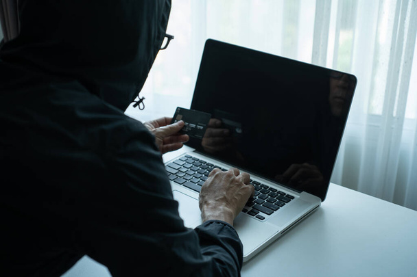 Rearside of man dressing black hood jacket holding credit card and checking password on labtop computer. hacker scammer concept - Foto, imagen