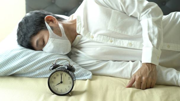 目覚まし時計でベッドの上で眠って外科マスクを身に着けているアジアのビジネスマン - 写真・画像