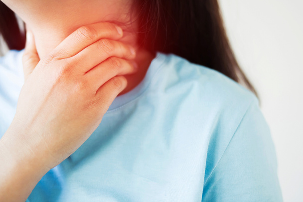 Боль в горле. Крупный план молодой женщины больной держа ее воспаленное горло с помощью рук, чтобы коснуться больной шеи в голубой рубашке на сером фоне. Медицинская и медицинская концепция. Фокусируйтесь на боли
. - Фото, изображение