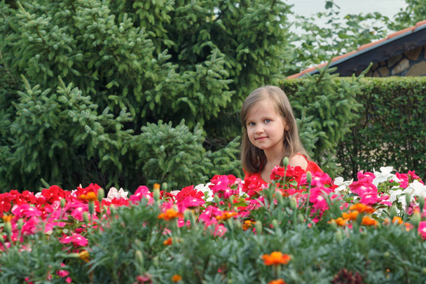 Kleines Mädchen mit langen blonden Haaren in rotem Kleid kauert hinter Blumen vor einem Hintergrund aus grünen Büschen und Tannen und schaut weg - Foto, Bild