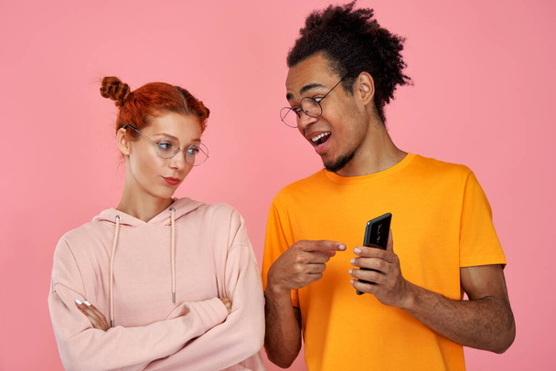 Satisfied black man points at screen on cell phone, demonstrates posted photos in social networks, browses on internet. Serious ginger woman looks at phone, excited with smartphone or app features - Fotografie, Obrázek