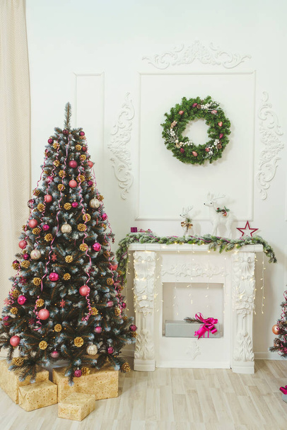 Két számjegyű fehér karácsonyi szarvas egy kandallón. Kandalló dekoráció stukkó karácsonyi szarvas és fenyő ágak. Golyók rózsaszín arany burgundi stílusban egy karácsonyfán közel a kandallóhoz - Fotó, kép