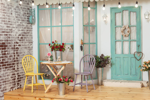 Τραπέζι κήπου και καρέκλες στην καλοκαιρινή βεράντα του σπιτιού. Θερινή βεράντα με ρετρό λάμπες γιρλάντες. Όμορφη βεράντα με ανοιξιάτικες διακοσμήσεις στο φωτογραφικό στούντιο - Φωτογραφία, εικόνα