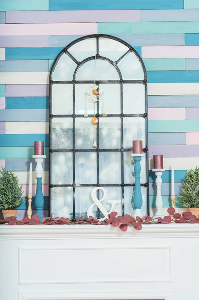 Holzbemalte Kerzenleuchter auf einem Kamin mit Spiegel. Frühlingsdekoration im Wohnzimmer. Spiegel in Form eines französischen Fensters im Innenraum vor dem Hintergrund einer mehrfarbigen Wand - Foto, Bild