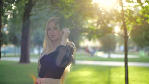 Young European lady with long chic light hair in sunset sunlight in a city park touches green leaves with her hand. Fashion model teenager in a stylish casual black blouse turns, shyly smiles, poses. - Footage, Video