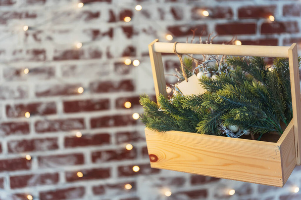 Luc ágak egy fadobozban egy téglafalon közelkép. Névtábla a szerszámosládán lucfenyő zöld ágakkal. Gyönyörű háttér karácsonyi díszek a háttérben bokeh koszorúk - Fotó, kép