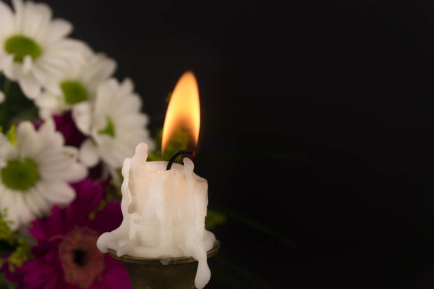 Σύντομη λευκό κερί καίει στο πάτωμα σε close-up μεταξύ των λουλουδιών σε σκούρο φόντο. - Φωτογραφία, εικόνα