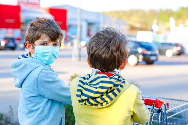 Two kids boys in medical mask as protection against pandemic coronavirus disease. Children using protective equipment against covid 19 and going for shopping in supermarket with cart trolley. - Photo, Image