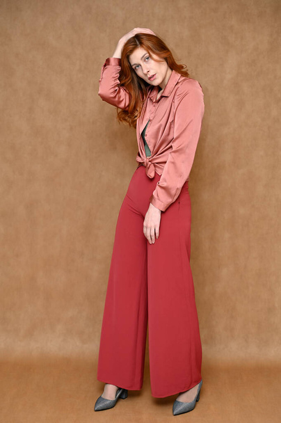 Full length vertical portrait of a posing standing straight Caucasian red hair cute young woman on a beige background. The model is dressed in a pink shirt and red pants, gray shoes. - Foto, Bild