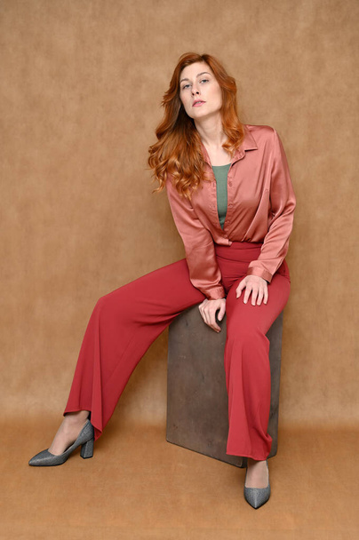 The model is wearing a pink shirt and red pants. Vertical portrait full length sitting on a stand Caucasian red hair pretty woman on a beige background. - Фото, изображение