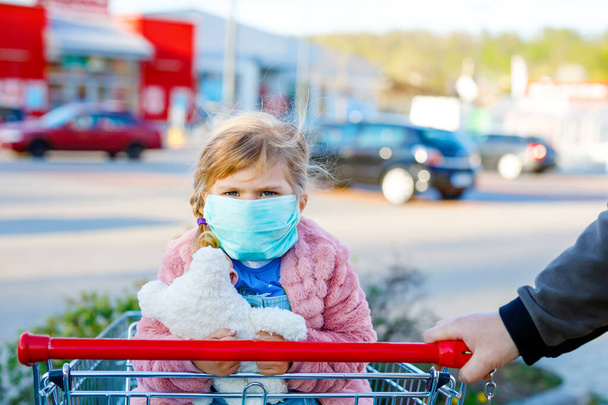Pikkutyttö lääketieteellisessä naamiossa suojana sepelvaltimotautipandemiaa vastaan. Söpö lapsi käyttää suojavarusteita covid 19 ja ostoksia supermarketissa ostoskorin työntää isä
 - Valokuva, kuva