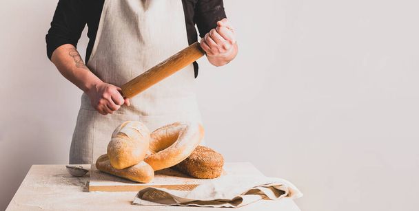 Puuvillaesiliinassa leipuri pitää puista neulaa. Erilaisia leipiä tuoretta leipää keittiön pöydällä
. - Valokuva, kuva