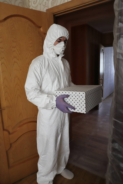 Ein Mann in Schutzanzug, Maske und Handschuhen mit einer Schachtel in der Hand brachte das Paket in die Wohnung zur Tür - Foto, Bild