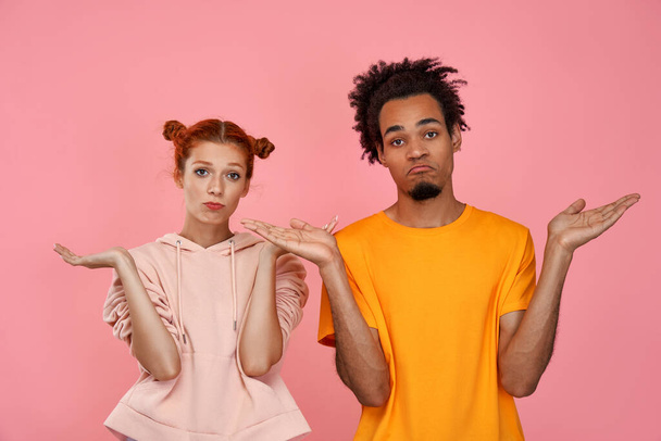 Photo of hesitant dark skinned boyfriend and ginger girlfriend look with uncertainty, receive suggestion, have doubtful facial expressions, isolated over pink background. Who cares, we dont know - Photo, Image