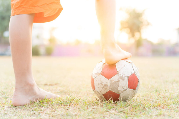 Τα πόδια ενός μικρού αγοριού με ένα παλιό κόκκινο και άσπρο ποδόσφαιρο το πρωί. Ένα μικρό αγόρι παίζει ποδόσφαιρο με θολό φόντο τη φύση - Φωτογραφία, εικόνα