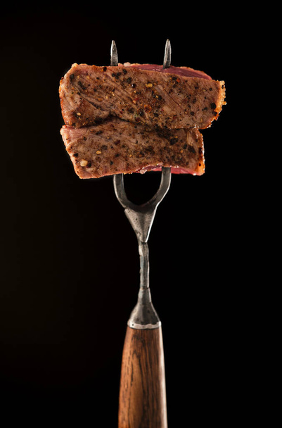 Egy szelet közepesen ritka grillezett steak Ribeye húsvillán, fekete alapon. Szaftos húsdarab villán. - Fotó, kép